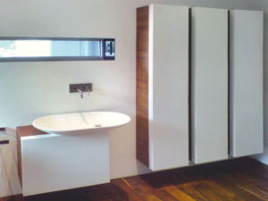 Badezimmer mit Holzmöbel und weißen Koffertüren. Im Innenraum der Türen sind kleine Regale angebracht. Das Waschbecken besticht durch seine moderne Form.